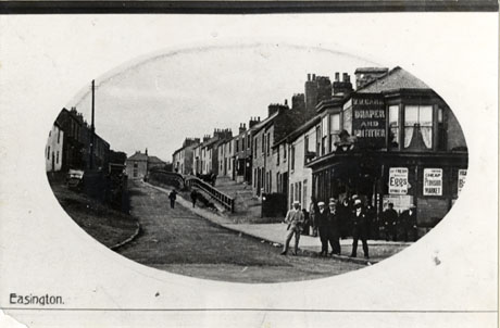 Photograph showing a street of terraced houses rising up a hill and set back from the road; at the corner of the right-hand side of the road at the end of the terrace is a shop, the ground floor windows of which are advertising fresh eggs and a Cheap Provision Market; on the first floor of the shop is an advertisement for a draper and outfitter; a group of what appear to be a dozen young men are standing on the pavement in front of the shop; the road is identified as Rosemary Lane