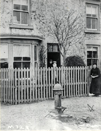 Photograph showing the front of a house with a bay window, in the doorway of which, a woman is standing holding an infant; an elderly woman is standing at the gate of the high fence, which surrounds the front garden of the house; in front of the fence in the street is a water pump; the photograph has been identified as being in Hall Walk, Easington