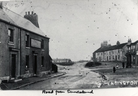 Photograph showing, close-up on the left-hand side of the picture the exterior of two terraced houses one of which may be a public house; in the far distance a row of low houses may be seen; in the middle of the picture is a road running away from the camera; on the right-hand side of the picture in the middle distance is a row of two-storied terraced houses; three figures, one of whom is a child, can be seen indistinctly;the photograph has been identified as Easington and Road From Sunderland