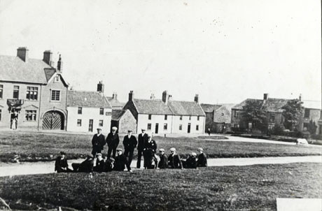 Photograph showing a group of fourteen men sitting on grass on the side of a road running horizontally across the picture; on the other side of the road is a further stretch of grass bordered by large building possibly a public house and two smaller houses; on the right of the grassed area are two or three substantial houses; figures can be seen indistinctly in the doorway of the large building; the photograph has been identified as The Village Green, Easington Village