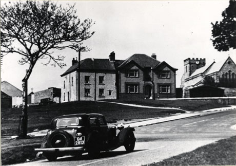 Photograph of a motor car, registration number GR 2813, driving up a road, with a large house, next to the church, on the far side of the road; the house has been identified as Hill House, Easington Village