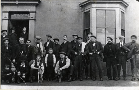 Photograph of seventeen men and three boys outside The Half Moon Public House, members of the Whippet Handicap Club; a man and a boy are each holding a whippet, and two women can be seen standing in the doorway of the public house where the name of the licensee, Matt. Cruddace, can be seen