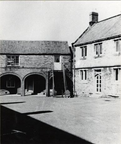 Photograph of the exterior of two wings of Leeholme Hospital and the courtyard; one wing has an arcade on the ground floor and a flight of steps to the first floor; the other wing has three windows on the first floor and two windows and a door on the ground floor