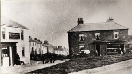 Photograph described as Masons Arms Corner showing the exterior of the front of The Masons' Arms with a street of houses behind it, and the front of a large detached house with a shop front in it; a few very indistinct people can be seen standing near the buildings