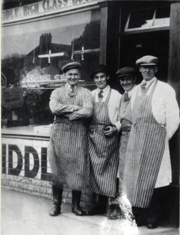 Photograph of four men standing outside Siddle's butcher's shop; each man is wearing a striped butcher's apron and are identified as from left to right Jack Mills; Burt Green; Ted Green; and Mr. Siddle second from the left; part of the shop window can be seen