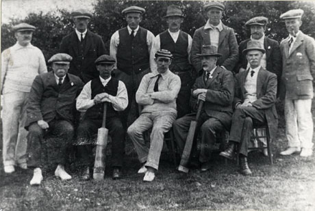 Photograph of the twelve members of the Easington Cricket Team in 1922; seven men are standing behind five men seated on chairs, two of whom are holding cricket bats; five of the men are wearing cricket whites and the rest ordinary suits and ties; the men are photographed against trees