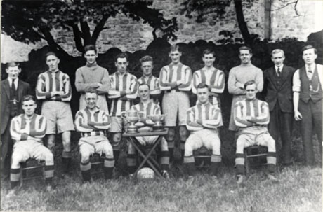 Photograph of the members of Easington Village Rovers Football Team in 1930-1931; the players are wearing the football strip and are accompanied by three men dressed in suits and ties; in front of the players is a table on which two trophies are placed; under the table is a football giving the year of the photograph