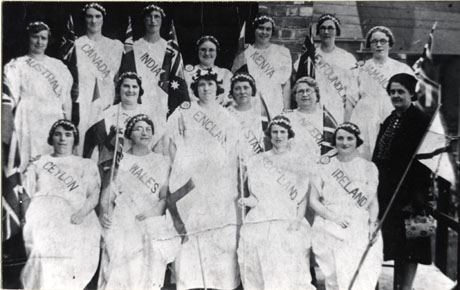 Photograph of members of Easington Village Women's Institute in a tableau representing nations of the British Empire; each lady is wearing a long and voluminous dress and has a sash across her chest with the name of a country on it , as follows: England; Scotland; Ireland; Wales; Ceylon; Nigeria; Malay States; Australia; Canada; India; Kenya; Newfoundland; Jamaica; each lady is holding the flag of the nation she represents; a lady in conventional dress is standing on the right-hand side of the photograph