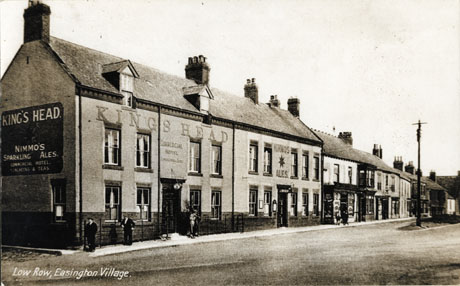 Postcard photograph entitled Low Row, Easington Village, showing the exterior of the King's Head public house and commercial hotel with the rest of the street of shops and houses in the distance; three indistinct figures of men can be seen outside the public house and two very small figures can be seen outside one of the shops