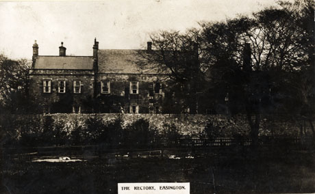 Postcard photograph entitled The Rectory, Easington, showing the exterior of the Rectory in the middle distance behind a wall; the roof, windows on the top floor and the tops of windows on the next floor can be seen; the foreground of the photograph is indistinct but it appears to be either a large garden or a park