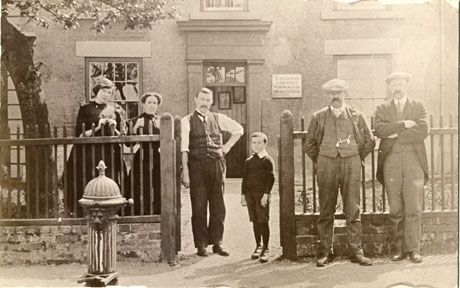 Photograph showing three men, two women, and a small boy, aged approximately seven years, standing near the fence in front of Easington and District Workmen's Club and Institute; the doorway and two windows of the club can be seen; in the foreground is the water pump