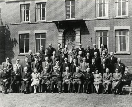 Photograph of the forty men and four women members of Easington Rural District Council posed outside a large brick building with an imposing entrance in stone; the photograph was taken in 1954, the sixtieth anniversary of the creation of urban and rural district councils; the man on the extreme right on the front row is Mr. Cain