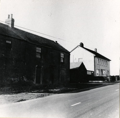 Photograph of the exterior of an old house with a newer house in the distance; two doorways and three windows can be seen in the older house, which is identified as Sandycar's Farm, Durham Road