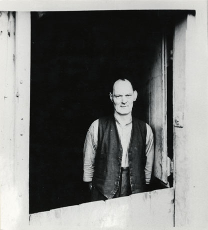 Photograph of the head and torso of a man looking through the open top half of a split door; he is wearing an open-necked shirt and an open waistcoat; nothing can be seen behind him in the interior of the building; he is identified as Mr. Tommy Hornsby, the last blacksmith in Easington Village
