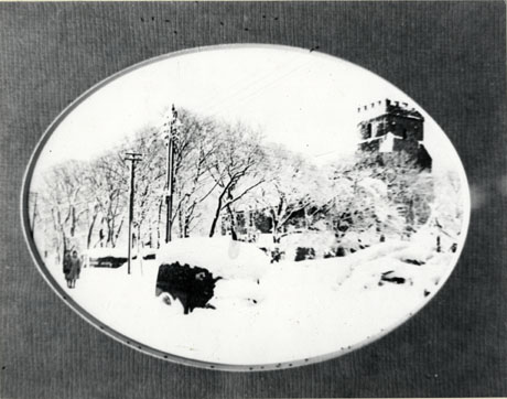 Photograph showing Easington church under snow with trees covered in snow and a vehicle, identified as Moore's delivery van, covered in snow; the scene is described as taking place during Easington's Worst Winter 1941; two small indistinct figures can be seen on the left-hand side of the picture