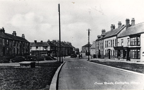 Postcard photograph entitled Cross Roads, Easington Village showing the view of the crossroads as in easv0044 taken from Low Row, showing the facades of shops and the King's Head on the right of the photograph and the facades of houses and shops further in the distance on the left of the photograph; the rear of a car and a horse-drawn vehicle can be sen at the crossroads