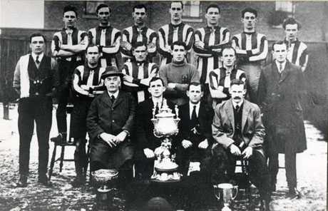 Photograph of the eleven members of the team of Easington Rovers Football Club and six men dressed in suits and ties; the group is posed in front of houses with what may be snow on the ground; the players are in the strip of the club and the men on the front row of the group have three trophies in front of them