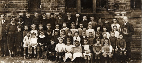 Photograph of a group of forty five children posed against a brick building with two men and one woman standing beside them; a child on the front row is holding a notice which reads Easington C.E. Group 4 30th August 1921; the children who appear to be approximately six years attend the Church of England School in Easington Village