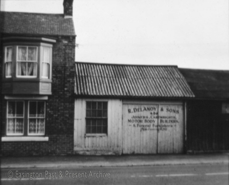 Photograph of the front of a house with bay windows on the first and second floors, with a wooden shed, with corrugated iron roof, attached; the doors of the shed bear the following words: R. Delanoy & Sons Joiners, Cartwrights, Motor Body Builders & Funeral Furnishers; the house and shed have been identified as being in Easington Village