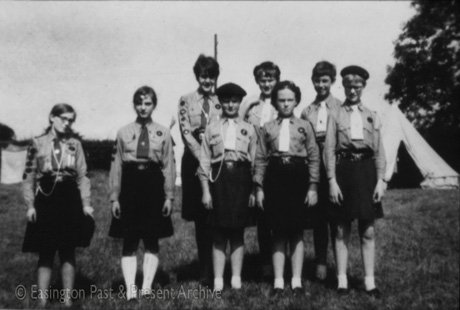 Photograph of eight girls, aged approximately between twelve and fourteen years, dressed as Girl Guides, standing in front of tents; they are wearing dark skirts, blouses with badges on them, ties, socks and shoes; two are wearing a lanyard as a badge of office; they have been identified as being from Easington Village