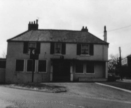 Photograph of an indistinct house with five windows on the ground floor and three on the first floor; it has a sign on a pole in front and been identified as The Half Moon at Easington Village