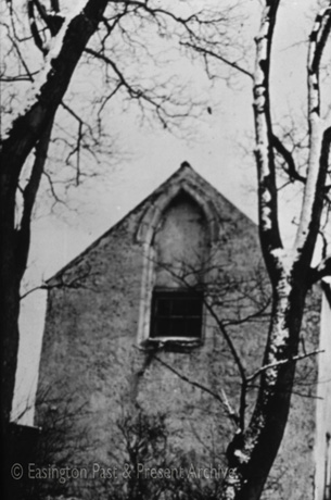 Photograph showing a pointed narrow window in the apex of a narrow gable, seen through the branches of a tree with snow on them; it has been identified as a tithe barn in Easington Village