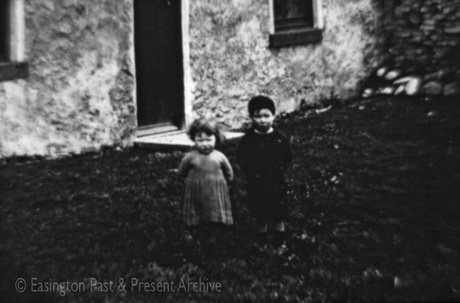 Photograph showing a small girl, aged approximately three years, wearing a dress, standing with a small boy, aged approximately five years, wearing a dark garment; they are both standing outside the doorway of a building the door and bottom of two windows of which only can be seen; they have been identified as Children At Moor Edge