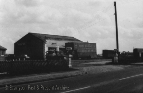 Photograph showing part of the front of a building with the word Wrights on it; in front of the building are a pantechnicon, with the words Ferens Transport Ltd. on it, and a lorry carrying crates; in the foreground is the surface of the road, a fence and gate posts to the garage on which Mobil is being advertised; the garage has been identified as being in Easington Village