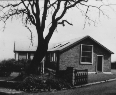 Photograph of the exterior of the end and front of a single-storey building with a window and door on the end and a porch on the front; a large tree is growing at the front of the building, which has been identified as The Mortuary, Easington Village