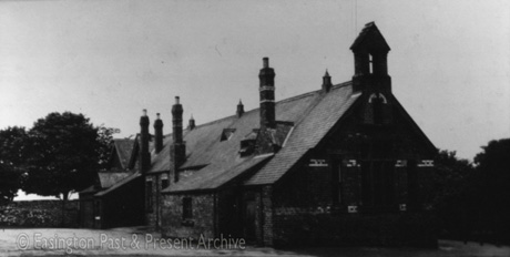 Photograph of the back and side of an indistinct single-storey building with three long chimney stacks and three ventilation cowls on the apex of the roof; there is a bell cote on the end of the building and what appear to be bands of coloured brick on its end wall; the building has been identified as Easington Village School