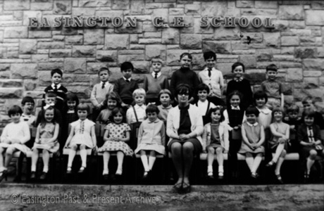 Photograph of twenty eight children, aged approximately six years, posed against a wall, on which the words Easington Church of England School are written; they are accompanied by a woman in dark glasses, presumably their teacher
