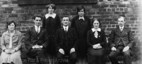 Photograph of three men and four women, posed against a brick wall; the men are wearing suits and the women dresses; they have been identified as teachers at Easington Village School