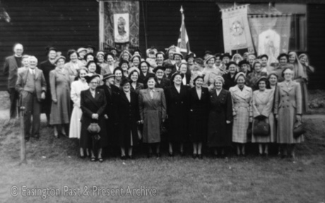 Photograph of approximately fifty three women, wearing overcoats, grouped with three banners and a Union Jack on a lawn; they are accompanied by a clergyman and another man; they have been identified as members of Easington Village Mothers' Union