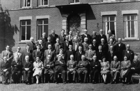 Photograph showing forty one men, wearing suits, and four women, wearing suits and overcoats, posed in four rows against the front of a large brick building with a doorway with a canopy and stone surround; they have been identified as Diamond Jubilee, Easington Village, which may indicate the Diamond Jubilee of Easington Urban District Council or of Easington Parish Council, both of which would have been established in 1894; the Diamond Jubilee of either organisation would have been 1954