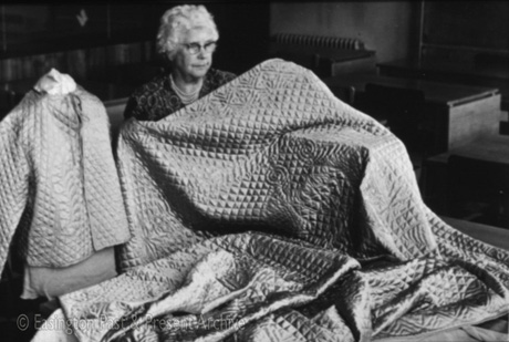 Photograph showing the head and shoulders of an elderly woman wearing glasses, pearls and a flowered dress, holding a quilt in front of her; on the left of the photograph is a dummy on which there is a quilted jacket; she has been identified as Emma Drydon of Easington Village and the quilting as Durham quilting