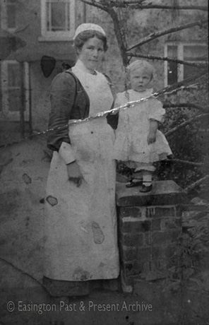 Photograph showing a woman dressed in an ankle-length apron, a dark dress. a white cap, and white false cuffs, standing beside the gate post of a garden wall on which a small boy, aged approximately two years, dressed in a frilly dress, frilly pantaloons. and dark shoes with straps, is standing; behind them a bush and the facade of a house can be seen indistinctly