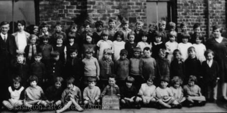 Photograph showing fifty one children, aged approximately between five years and seven years, posed in four rows against the wall of a brick building; there are a man and a woman standing on the left and a woman on the right; a child on the front row is holding a notice reading Easington Church of England School Infants 1929; the man and the women are among those portrayed on easv0071