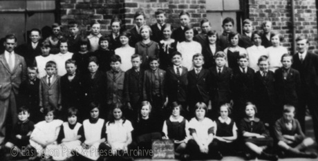 Photograph showing forty eight children, aged approximately between seven and ten years, posed in four rows against a brick building; a man is standing either side of the group; a child on the front row is holding a notice reading: Easington Village Church of England School......1929; the men are two of the men in easv0071