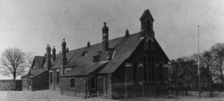Photograph showing the side and rear of a single-storey building with three tall chimney stacks and three ventilation cowls along the apex of its roof; it has bands of coloured brick along its side wall; the view is the same as that in easv0066, which was taken in summer, whereas this photograph was taken in winter and the facade of a house across the road from the building can be seen; the building has been identified as Easington Old Village School