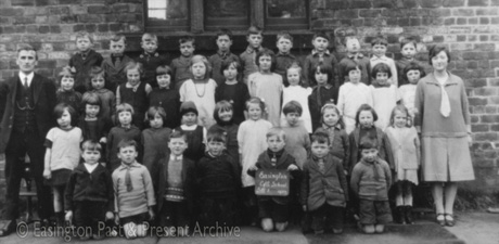 Photograph showing forty one children, aged approximately seven years, posed in five rows against the wall of a brick building; a child on the front row is holding a notice reading: Easington Church of England School Standard 1 1927; there are a man and a woman standing with them, both of whom are in the photograph easv0071