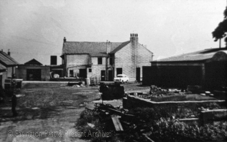 Photograph showing the rear of a two-storey stone building with the front of outhouses on the left of the picture; on the right is a large shed, and indistinct items are lying in the square of space between the house and its outhouses; a car is parked near the house, which has been identified as Cadwell House, Easington Village