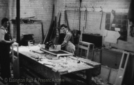 Photograph of a man standing at a bench covered in what appear to be shavings and pieces of wood; he is operating a machine; behind him are brick wall with tools hanging from hooks, planks of wood, cupboards cases and other containers; in front of the bench is another man standing holding what appears to be a wooden frame; the photograph has been identified as Inside Cadwell House - Ronnie Pritchard and Neil Summers