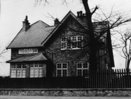 Photograph showing the side facing the road of a brick house; it has a large gable on the side facing the road and on the side away from the road; on the side facing the road are two long windows, a wide window, a small narrow window, and two bay windows; the building has been identified as The Rectory, Easington Village
