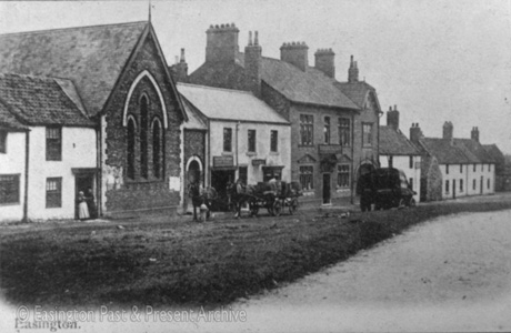 Postcard photograph entitled Easington, showing, on the left, the facade of a cottage at the door of which two women can be seen; next to that is the end of a chapel and next to that the facade of what may be a public house; next to that is a larger building, also with a sign above its doorway; beyond that building are cottages; there is brewer's horse-drawn dray in front of the public house and a cart standing on the verge; the picture has been identified as Long Row in Easington Village