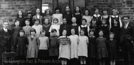 Photograph showing thirty two girls, aged approximately between eleven and thirteen years, posed in three rows against the wall of a brick building; a girl on the front row is holding a notice reading: Easington Church of England School Senior Girls 1927; two men, the one on the left of whom is featured on easv0071, are standing with the girls