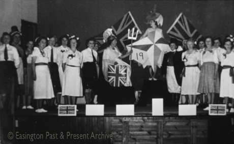 Photograph showing approximately fifteen women standing on a stage wearing costumes as Welsh women, Scots in kilts, and a man wearing at top hat and frock coat; five of the women are wearing light-coloured dresses with sashes and flowers pinned to their frocks; in the centre is a woman dressed as Britannia, with a trident and a shield with a Union Jack on it, and woman wearing a helmet and holding the symbol of the Festival of Britain in 1951; they have been identified as members of the Women's Institute in Easington Village