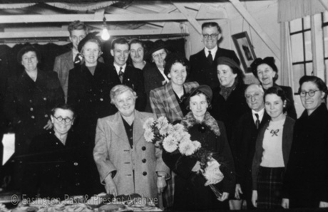 Photograph showing a group of ten women and six men standing beside a table on which there are plates of cake and biscuits; most of the people are wearing overcoats and a woman at the front of the group is holding a bunch of chrysanthemums; the ceiling of the room in which they are standing has low beams on which paper streamers can be seen; the photograph has been described as Decorating Church Hall, Easington Village