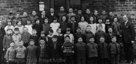 Photograph showing forty eight children, aged approximately seven years, posed in four rows against a brick wall; a man is standing either side of the group; a child on the front row is holding a notice reading: Easington Church of England School Standard 3-4 1927