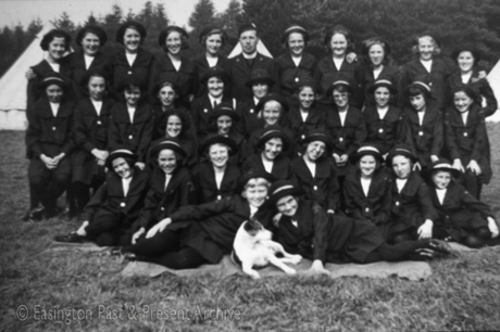 Photograph of thirty four girls wearing a uniform of dark skirt, dark jacket, dark stockings and dark hat, posed in five rows with trees and tents behind them; they are accompanied by, in the middle of the back row, a clergyman in uniform, and, on the front row, a small white dog; they have been described as members of the Methodist Girls' Brigade from Easington Village