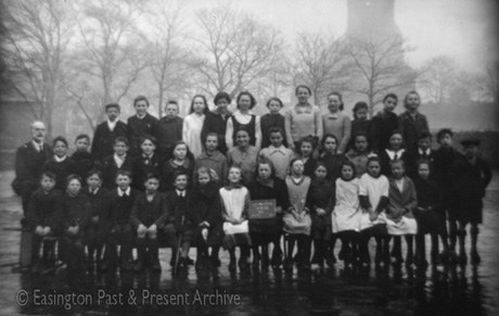 Photograph showing forty one children, aged approximately ten years, posed with leafless trees and the end of a church behind them; a man is accompanying them; they have been identified as pupils at Easington Village School